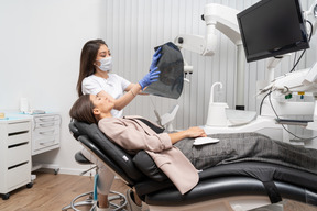 Женщина-стоматолог в полный рост и ее пациентка смотрит на рентгеновский снимок
