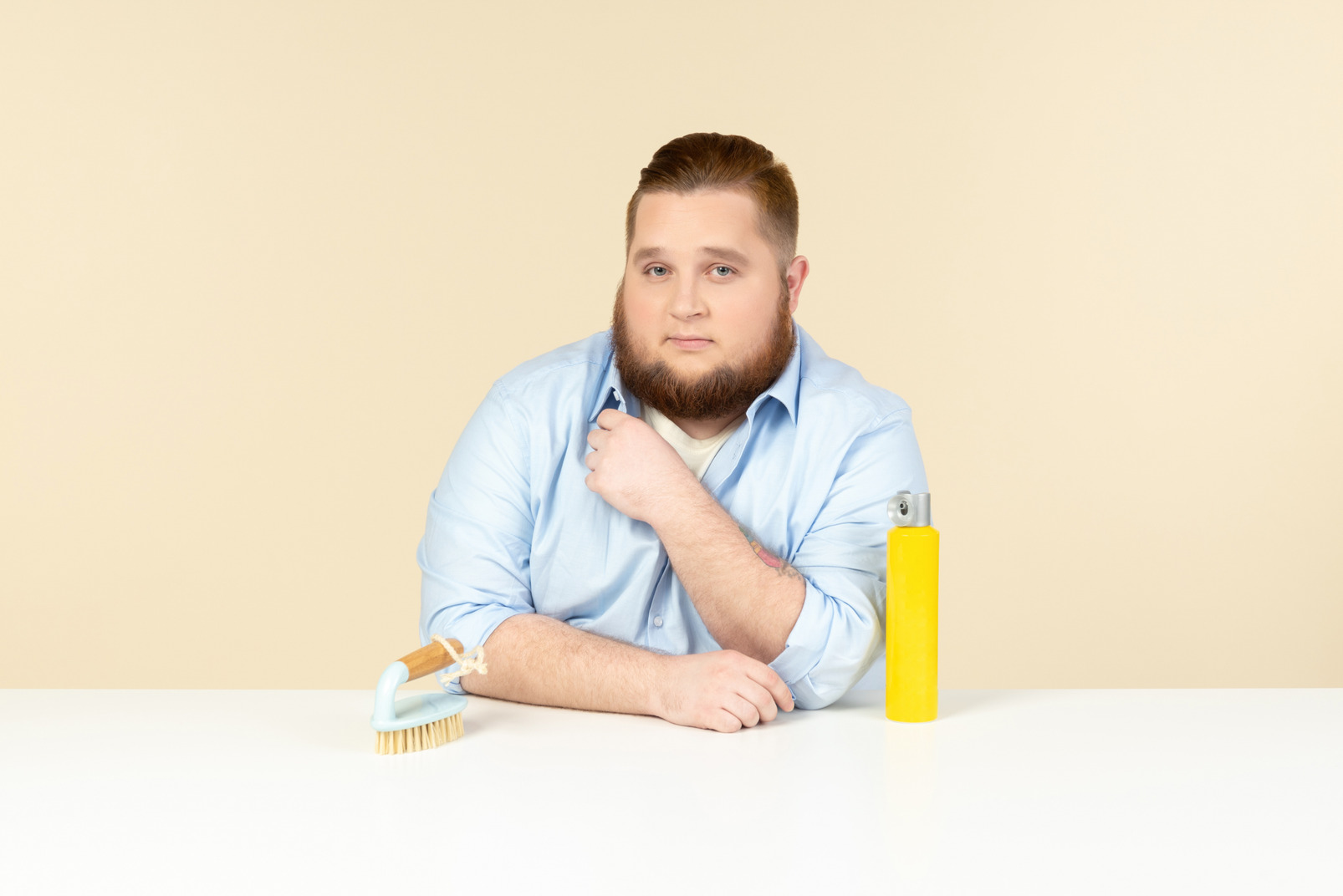 Serious looking young overweight man sitting at the table with cleaning spray and brush on it