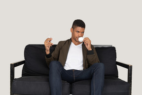 Front view of young man sitting on a sofa and holding cigarette and drinking coffee