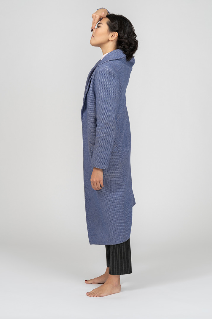 Young asian woman in long blue coat standing in profile and touching her nose with the hand