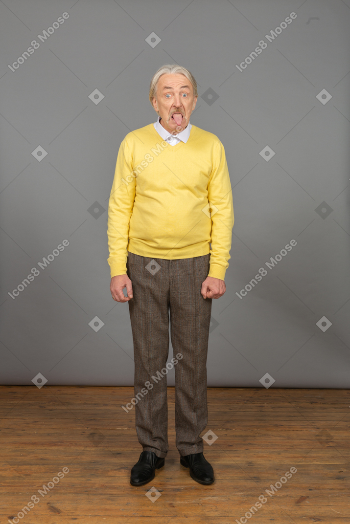 Vista frontal de un anciano curioso en jersey amarillo mostrando la lengua y mirando a la cámara