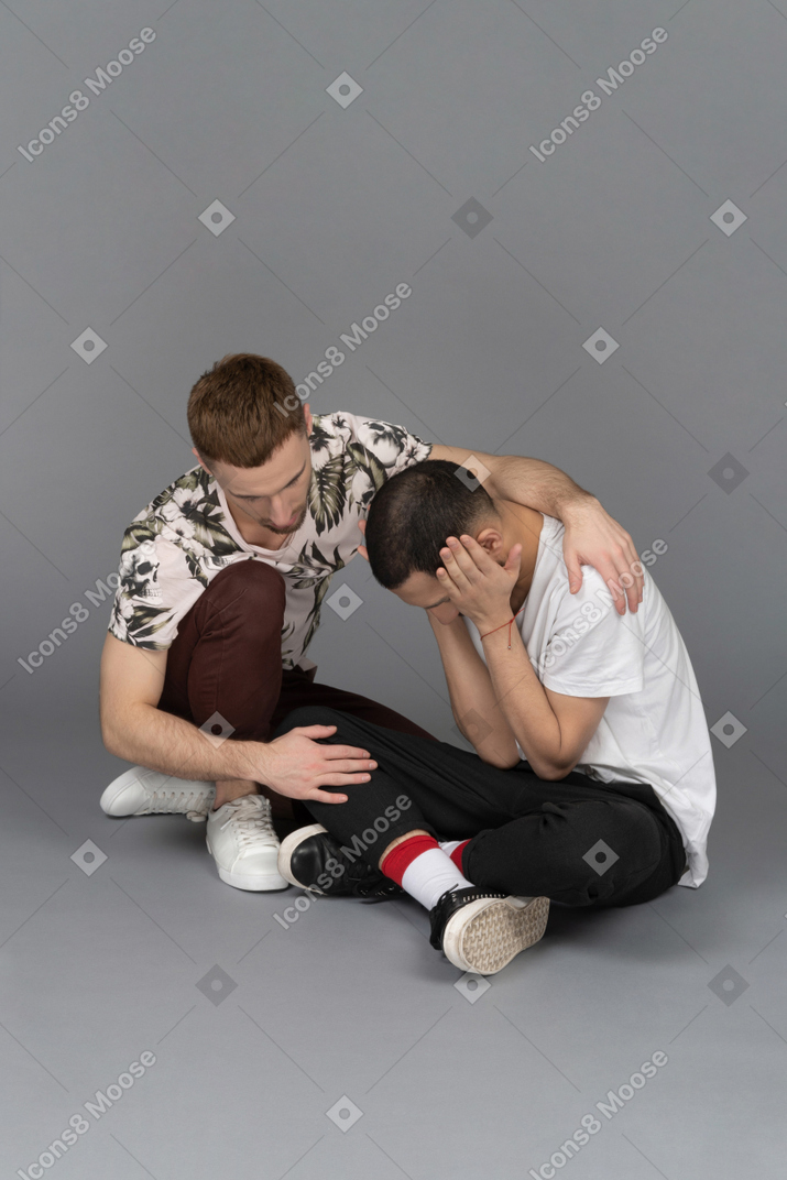 Young man sitting on the floor and comforting another distressed young man