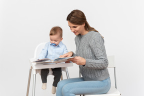 Young mom reading a book to her little son sitting in a highchair