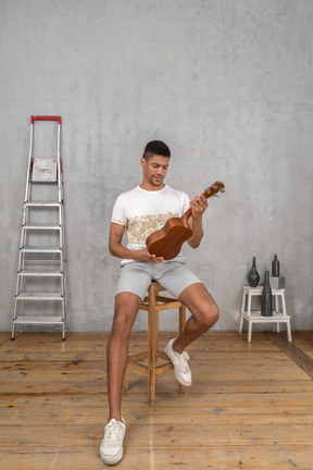 Vista frontal de um homem em um banquinho examinando um ukulele com cuidado