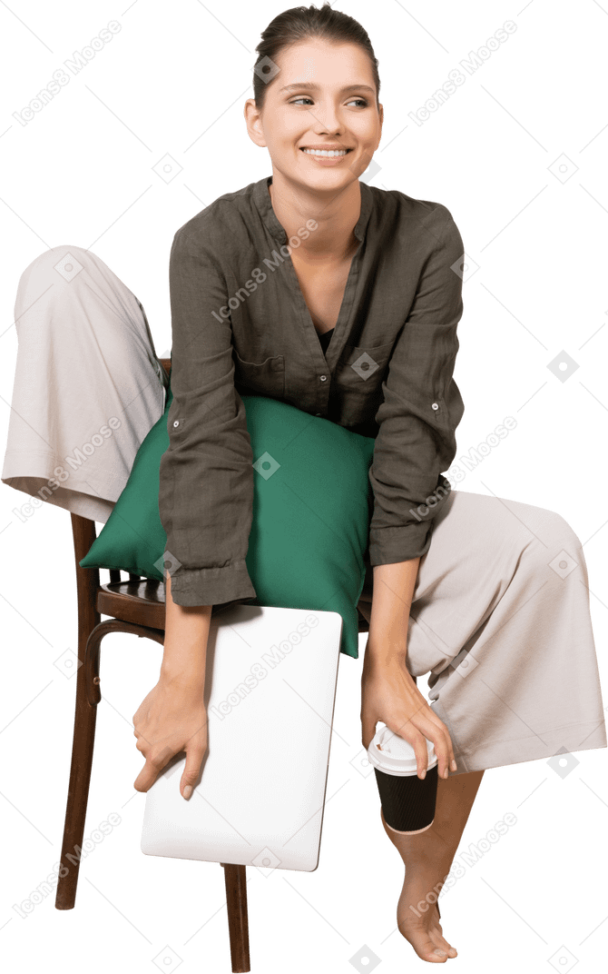 Vista frontale di una giovane donna sorridente seduta su una sedia e tenendo il suo laptop e toccando la tazza di caffè