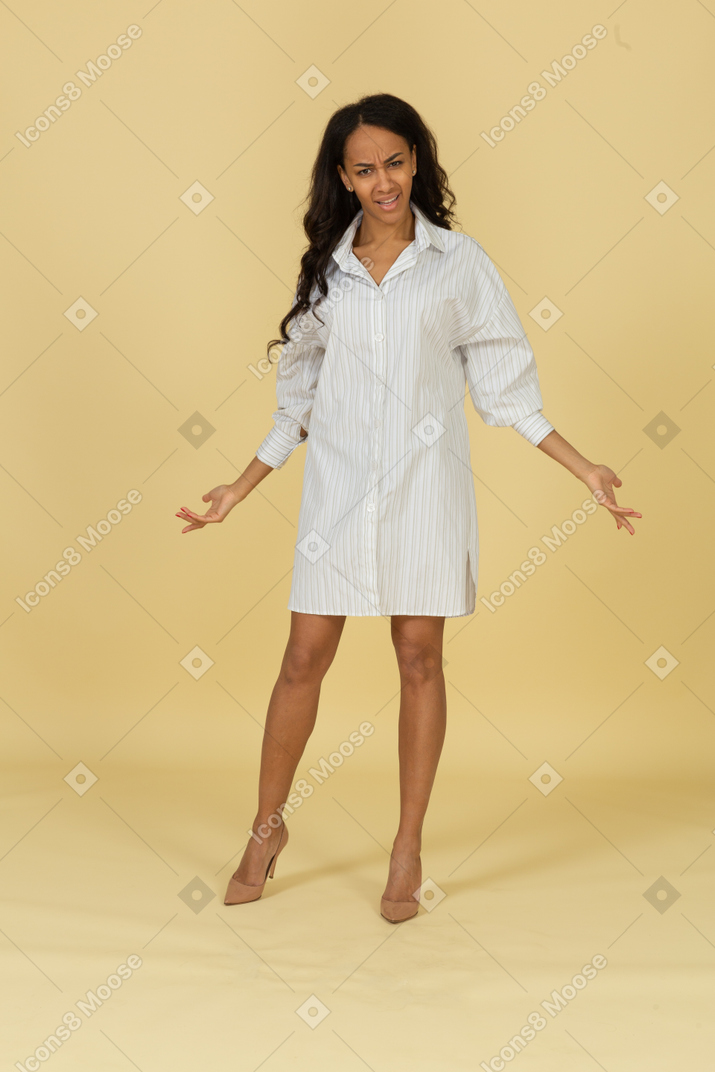 Front view of a questioning dark-skinned young female in white dress outspreading hands