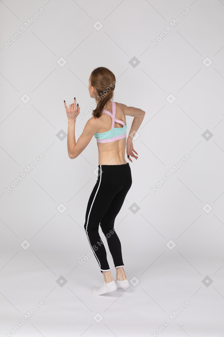Vista posterior de tres cuartos de una jovencita en ropa deportiva levantando las manos mientras baila