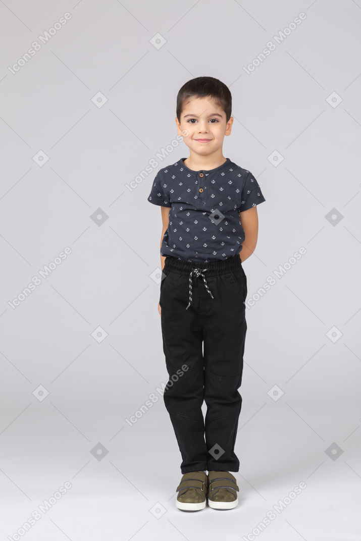 Vista frontal de um menino feliz em roupas casuais em pé com as mãos atrás das costas e olhando para a câmera