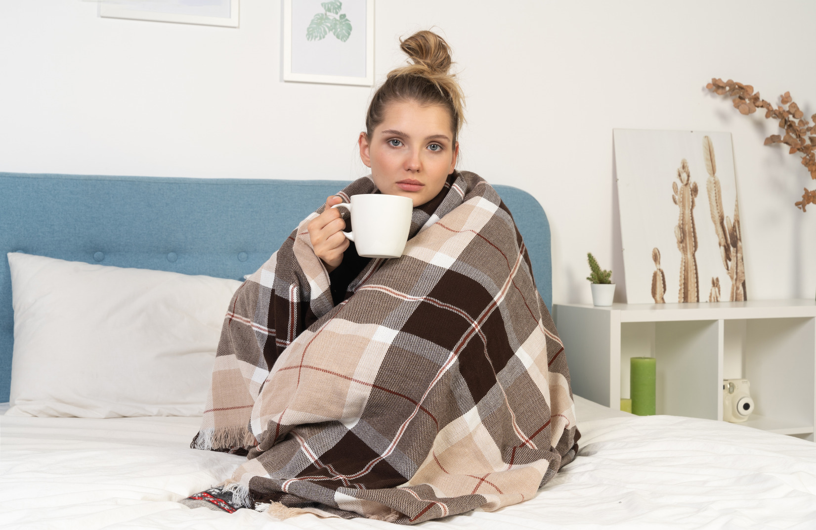 Front view of an ill young lady wrapped in checked blanket in bed holding a cup of tea