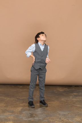 Вид спереди мальчика в сером костюме, позирующего с рукой на бедре и смотрящего вверх