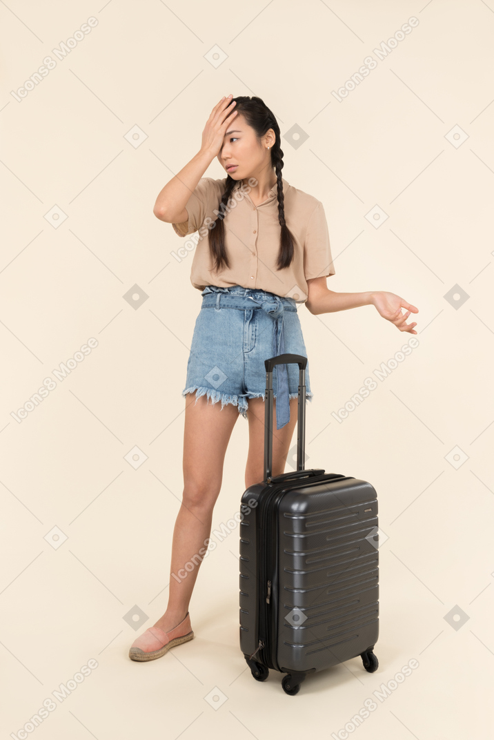 Young female traveller standing near suitcase and closing her face with a hand