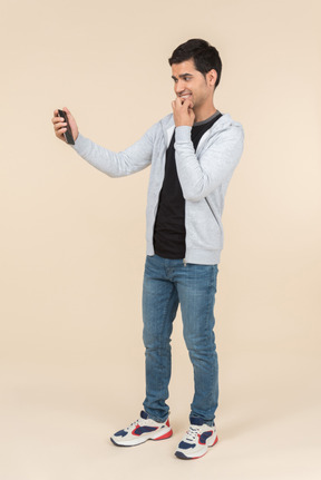 Young caucasian guy holding smartphone and bitting his fist