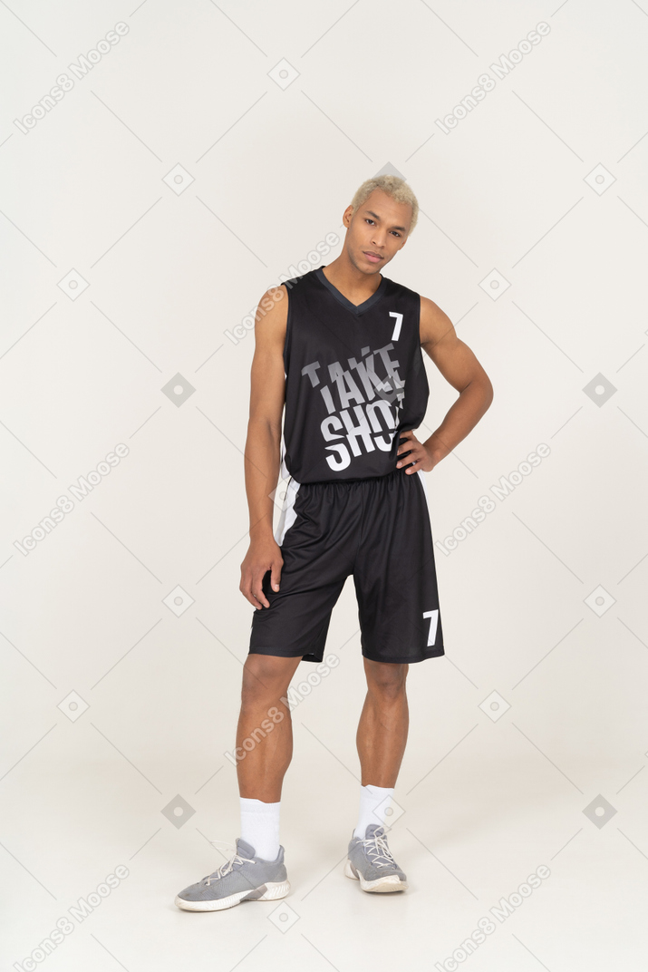 Vista frontal de un joven jugador de baloncesto masculino poniendo la mano en la cadera y mirando a la cámara