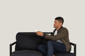 Dreiviertelansicht eines jungen träumenden mannes, der mit einer tasse kaffee auf einem sofa sitzt
