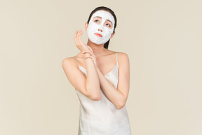 Rêveuse jeune femme asiatique avec masque facial, touchant sa main