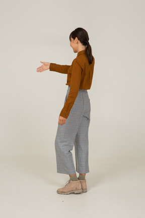 Three-quarter back view of a greeting young asian female in breeches and blouse outstretching hand