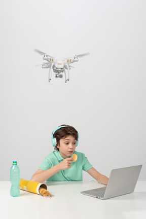 A boy in earphones is so engrossed in his laptop that he doesn't notice a hovering drone