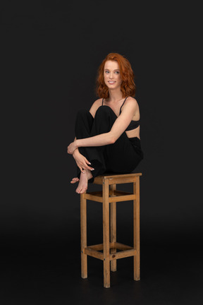 A three-quarter frontal view of the beautiful woman dressed in black pants and bra, sitting on the wooden chair and hugging her knees