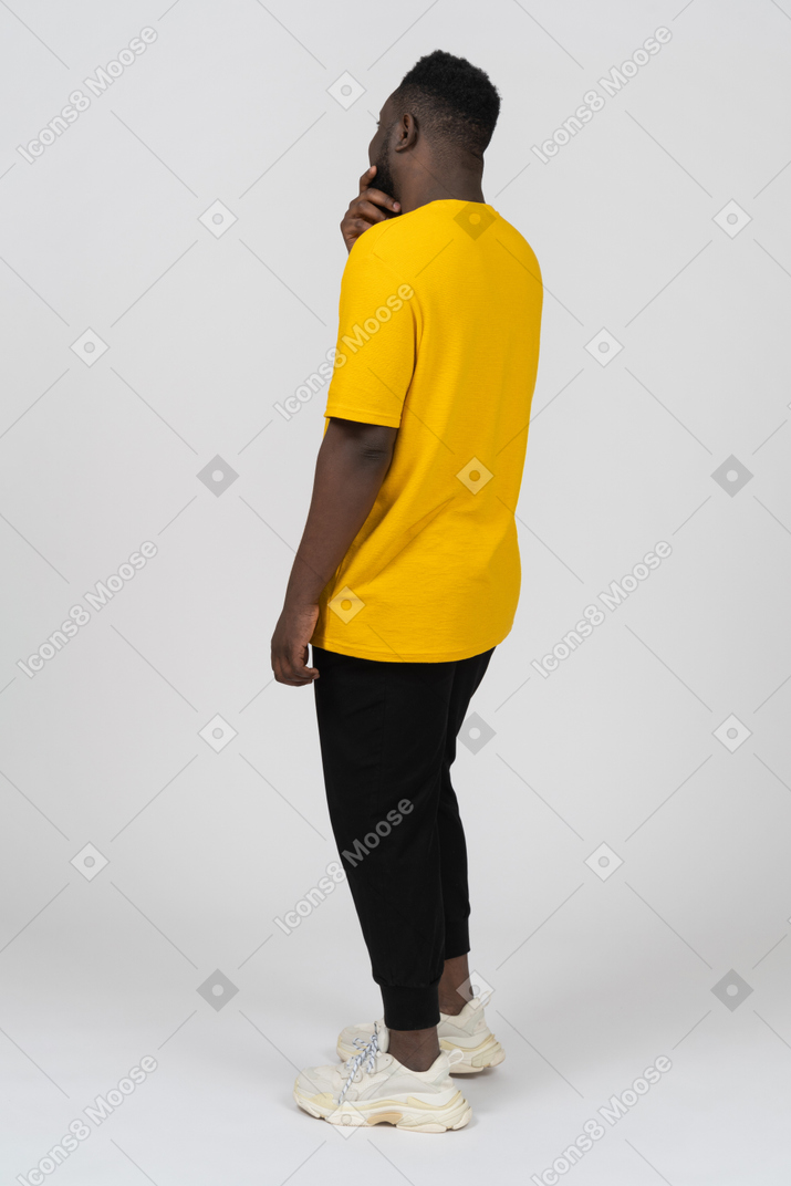 Three-quarter back view of a guessing young dark-skinned man in yellow t-shirt touching chin