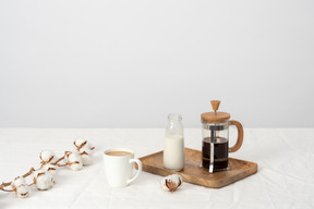 Coffee in french press and bottle of milk on wooden tray, large cup of coffee and cotton branch on the table