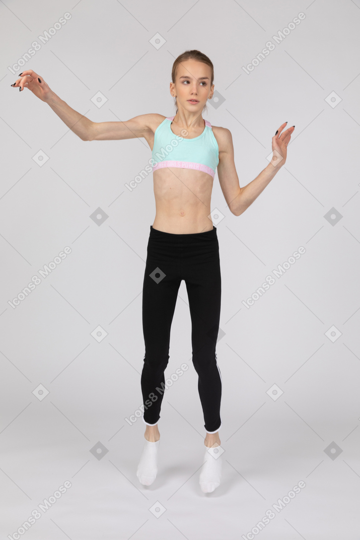 Vista frontal de una jovencita en ropa deportiva levantando la mano y mirando a un lado mientras salta