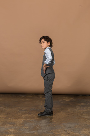 Vue latérale d'un garçon mignon en costume gris debout avec les mains sur les hanches et levant les yeux