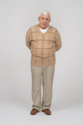 Front view of an old man in casual clothes standing with hands behind back and looking at camera