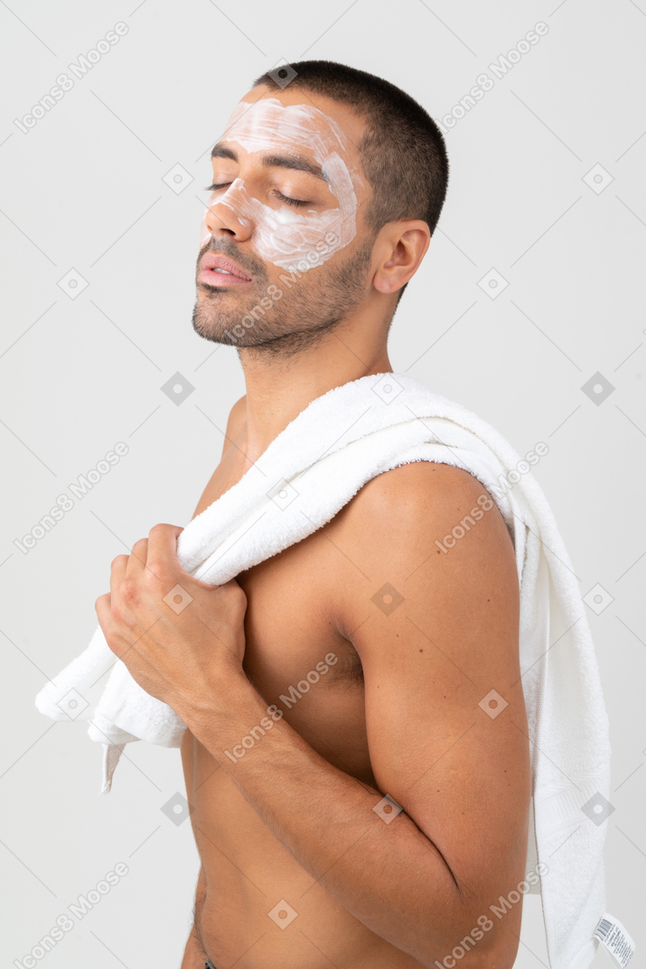 A topless man with eyes closed and moisturizer on his face holding a towel