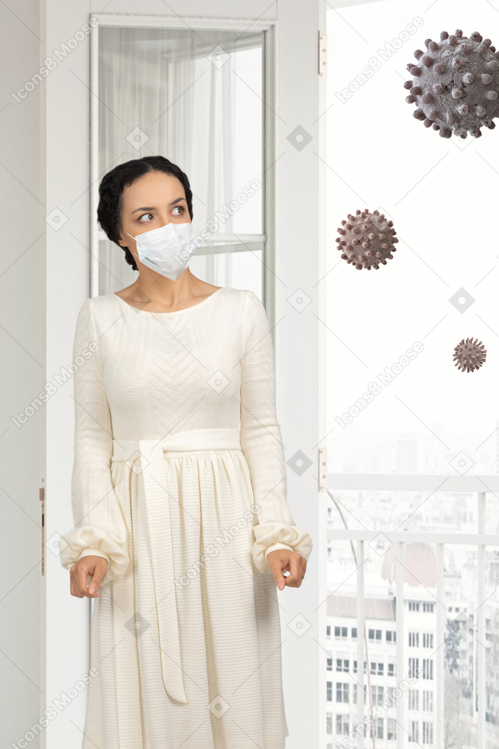 A woman in face mask standing next to a window with coronavirus floating in the air outiside