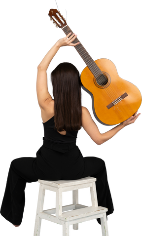 Back view of a young lady in black suit holding the guitar over head and sitting on stool