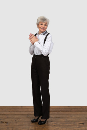 Vista di tre quarti di una donna anziana sorpresa vestita in abiti da ufficio mettendo le mani insieme guardando la fotocamera e sorridente