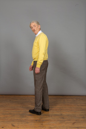 Vista lateral de um velho surpreso de blusa amarela, curvando-se, olhando para a câmera e fazendo caretas