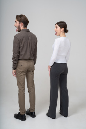 Vista posterior de tres cuartos de una joven pareja loca en ropa de oficina mostrando la lengua