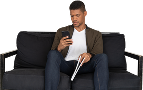 Front view of young man sitting on a sofa holding a magazine while using phone