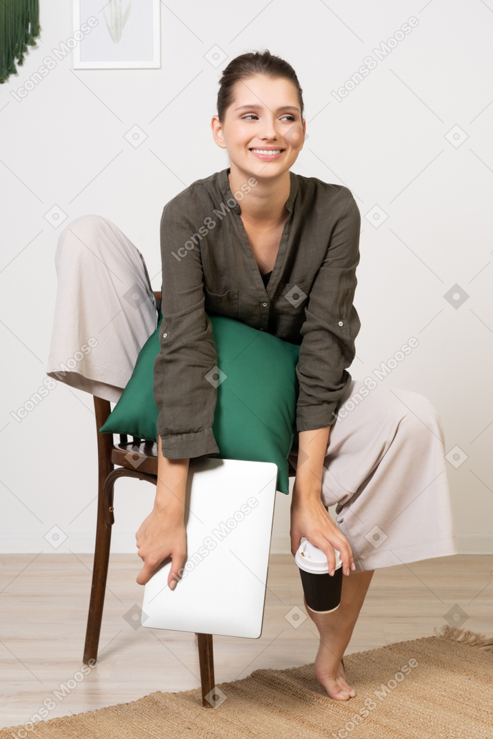 의자에 앉아 노트북을 들고 커피잔을 만지고 있는 웃고 있는 젊은 여성의 전면 모습