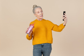 Old woman holding plastic cup to go and making selfie