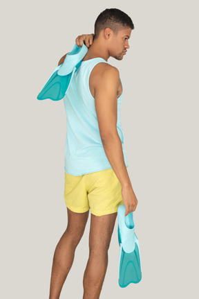 Side view of a man in yellow shorts and a blue tank top holding flippers