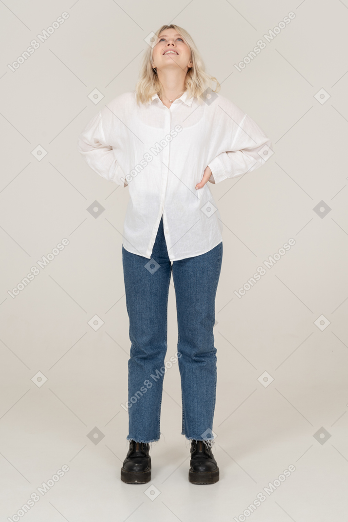 Front view of a blonde female in casual clothes putting hands on hips and looking up while smiling
