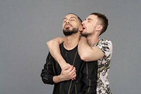Молодой кавказский мужчина обнимает другого мужчину со спины, переплетая руки и кусая ухо