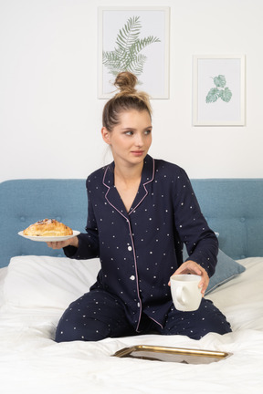 Vista frontal de una joven en pijama sosteniendo una taza de café y algunos pasteles sentado en la cama