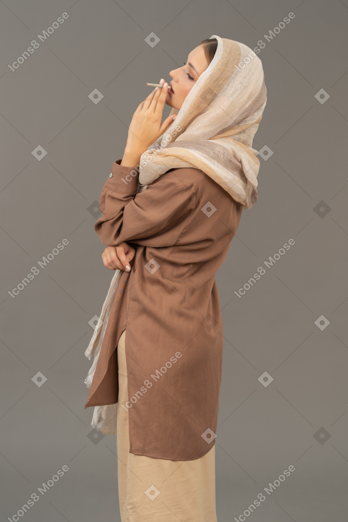 Side view of a young woman in headscarf and beige clothes smoking a cigarette