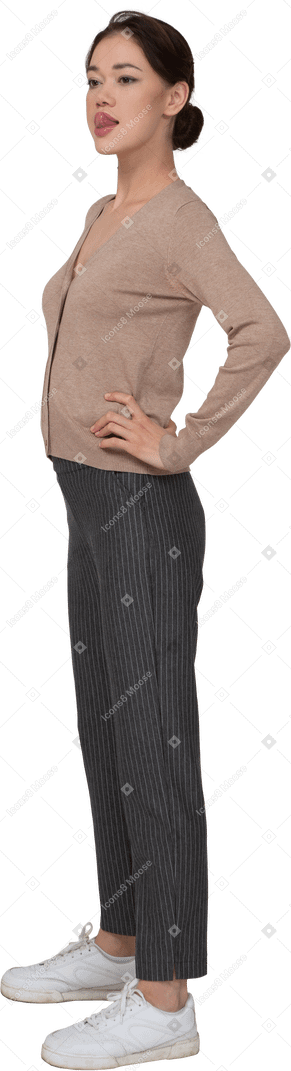 Vista di tre quarti di una giovane donna in pullover e pantaloni che mette le mani sui fianchi e mostra la lingua