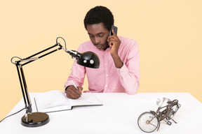 Apuesto joven escribiendo algo en un cuaderno y hablando por teléfono