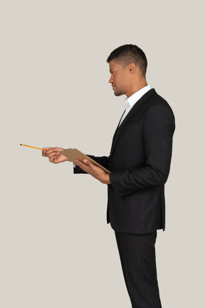 Standing in profile young man in black suit holding folder and pencil