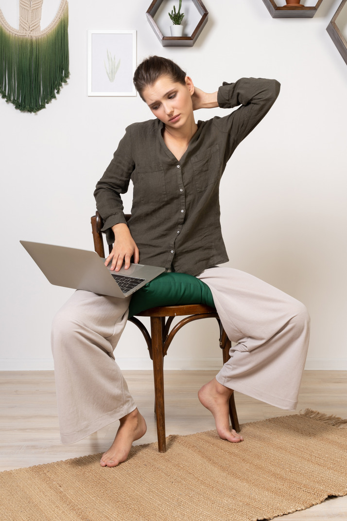 Front view of a tired young woman wearing home clothes sitting on a chair with a laptop