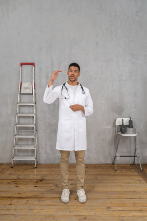 Front view of a young doctor standing in a room with ladder and chair showing a size of something