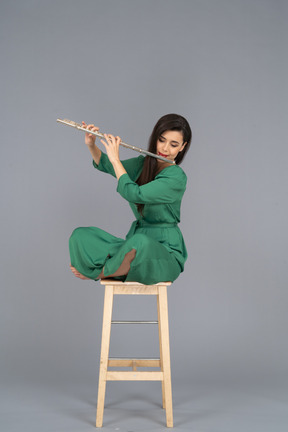Toute la longueur d'une jeune femme jouant de la clarinette assise, les jambes croisées sur une chaise en bois