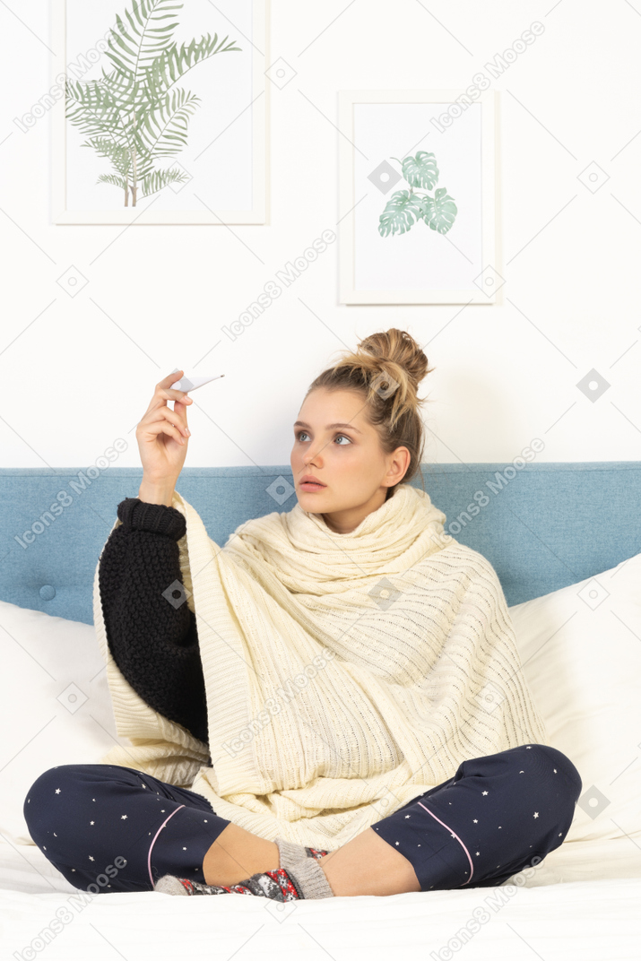 Front view of a young lady wrapped in white blanket sitting in bed with thermometer