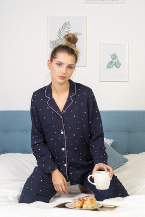 Front view of a young lady in pajamas holding a cup of coffee and some pastries on a tray