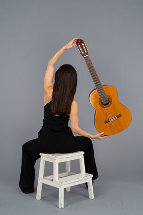 Vista posterior de una joven en traje negro sosteniendo la guitarra sobre la cabeza y sentada en un taburete